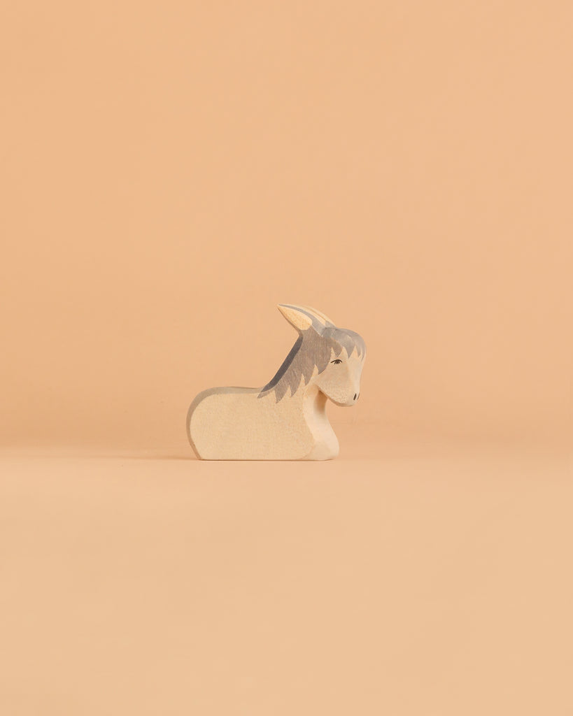 A small, minimalist goat figurine from the Ostheimer Handmade Wooden Farm Animals Set, handcrafted from sustainably sourced wood with grey and white accents, sits on a light peach background.
