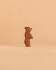 A small, brown bear figurine from the Ostheimer Handmade Wooden Woodland Animals Set stands upright on a plain beige background, embodying the charm and quality of handcrafted toys.