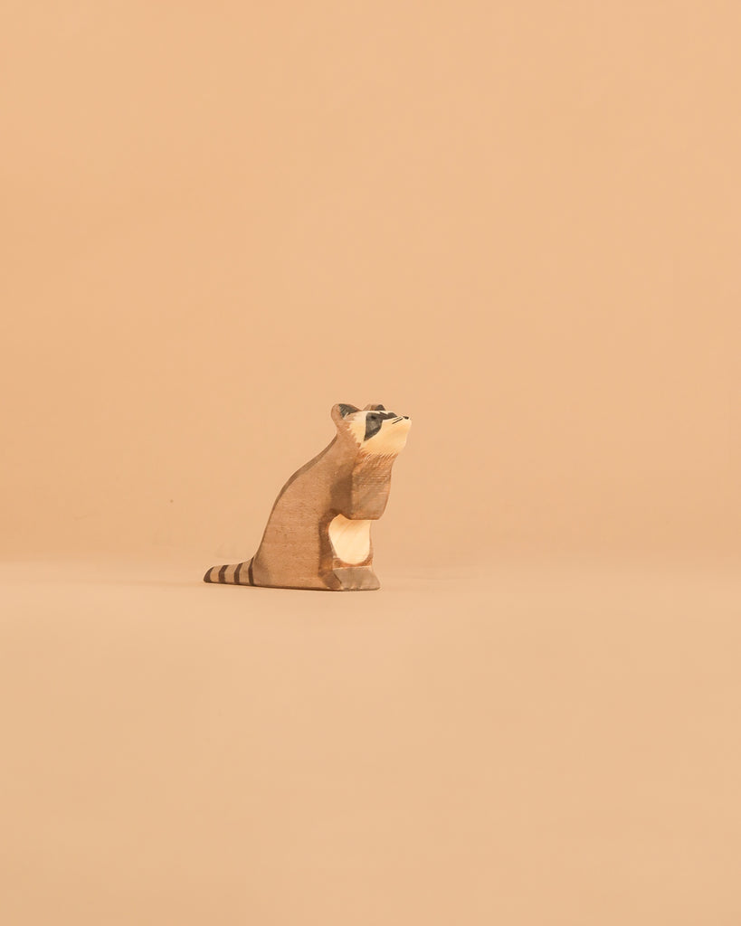 A small, carved wooden raccoon figurine from the Ostheimer Handmade Wooden Woodland Animals Set sits on a smooth beige surface with a matching beige background, reminiscent of handcrafted toys.