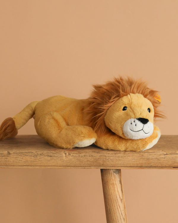 The Steiff Liam Lion Stuffed Animal, featuring a brown mane and tail tip, is shown lying on a wooden surface against a plain beige background.