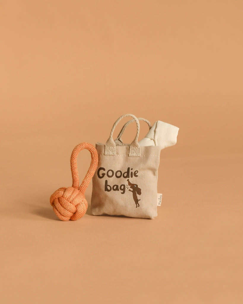 The Maileg Pet Supply, Toys—a small beige tote labeled "Goodie bag" with a dog silhouette and puppy accessories—stands beside an orange rope dog toy on a plain tan background.
