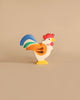 A Holztiger Rooster, made in Europe and painted in bright colors, features a red comb, yellow beak, white body with orange feathers, and a blue and green tail. This handcrafted wood piece stands upright on a neutral tan background and epitomizes the charm of HOLZTIGER figures.