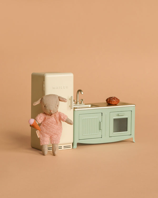 A small stuffed animal in a pink outfit holds an ice cream cone beside the Maileg Little Lambs Kitchen Set and Maileg Metal Fridge, with a pastry on the counter. The kitchen features a sink and oven against a beige background.
