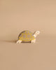A small, handcrafted wooden toy turtle placed on a beige background. The Holztiger Tortoise, reminiscent of classic HOLZTIGER figures, has a white head and legs with a soft grey shell accented by yellow grid patterns. It is carefully crafted from maple and beech wood.