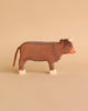 A wooden toy cow with a brown body, white hooves, a red nose, and tiny horns set against a beige background. Handcrafted out of maple and beech wood, this simplistic yet charming Holztiger Highland Cattle, Standing figure boasts minimal detailing for a rustic appearance. Made in Europe to ensure high quality craftsmanship.