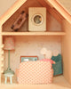 Experience the charm of the Maileg Fully Furnished Farmhouse Set + Surprise Gift, featuring a cozy room with a mouse doll relaxing on a pink polka dot sofa, surrounded by adorable dollhouse accessories—including a miniature TV, lamp, washing machine, and suitcase—all beautifully arranged against pastel wallpaper.