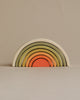 A set of colorful wooden nesting arches forming a rainbow, arranged from largest to smallest with non-toxic paint in muted colors including green, yellow, and orange, against a plain beige background.