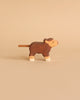 A small wooden toy cow with a rope tail is depicted against a beige background. Made in Europe, the Holztiger Highland Cattle, Small figure is brown with white accents on its legs and face, featuring a simple, hand-painted design.