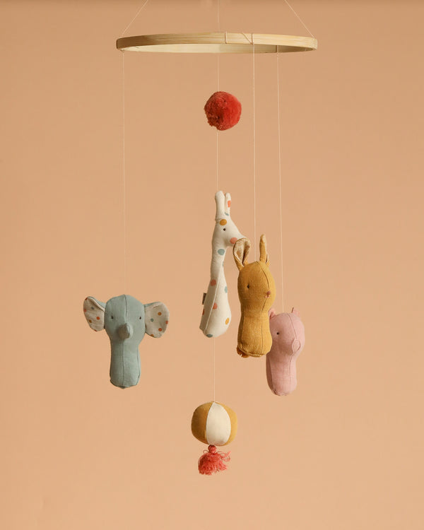 The Maileg Lullaby Friends Mobile hangs against a beige background, showcasing soft fabrics and unique prints: a blue elephant, spotted giraffe, yellow bunny, and pink bear suspended from strings with decorative balls above and below.
