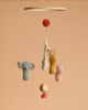The Maileg Lullaby Friends Mobile hangs against a beige background, showcasing soft fabrics and unique prints: a blue elephant, spotted giraffe, yellow bunny, and pink bear suspended from strings with decorative balls above and below.