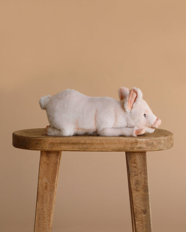 The Pig Stuffed Animal 11" lies on its side on a simple wooden stool, set against a plain beige background, showcasing its realistic plush design.