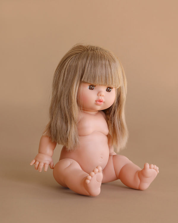 The Minikane Baby Doll (13") - Nina, featuring long, straight blonde hair and bangs, sits upright against a plain beige background. Clothing sold separately.
