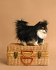 A Pomeranian Dog Stuffed Animal, featuring lifelike detail, stands on a closed wicker suitcase against a plain light brown background.