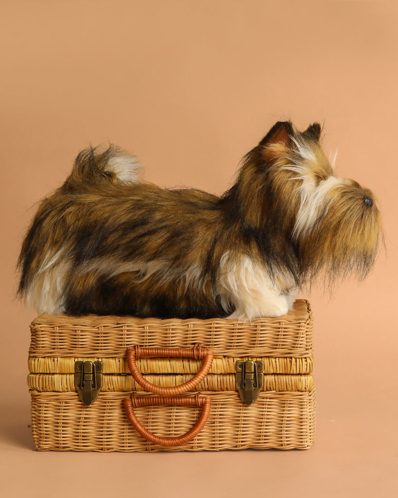 The Terrier Dog Stuffed Animal, a plush toy with brown and black fur, sits atop a closed woven wicker suitcase with tan handles, set against a plain beige background.
