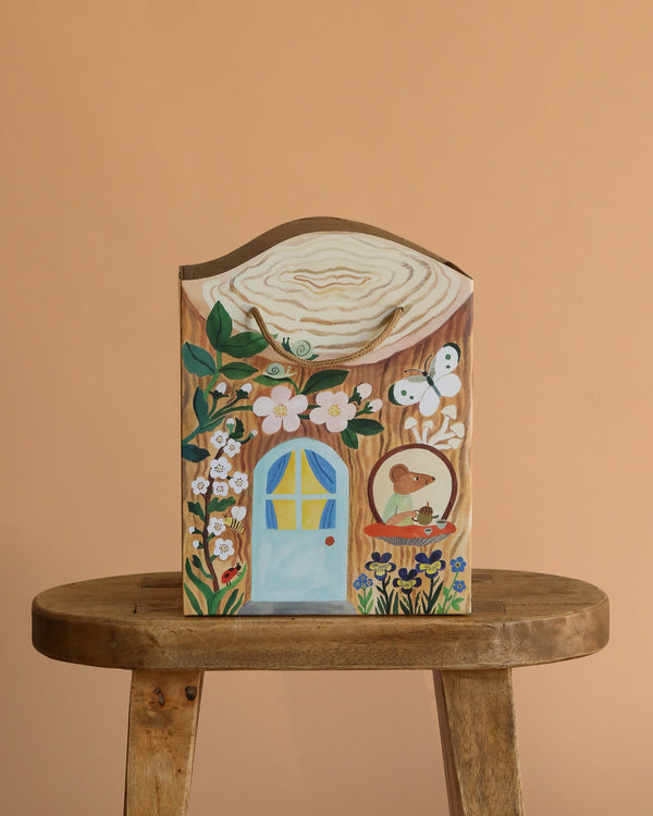 The Odin Parker x Gemma Koomen Gift Bag, adorned with a blue door, flowers, butterfly, and bird, rests on a rustic wooden stool against a peach backdrop, capturing cozy cottagecore charm.