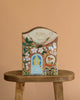 The Odin Parker x Gemma Koomen Gift Bag, adorned with a blue door, flowers, butterfly, and bird, rests on a rustic wooden stool against a peach backdrop, capturing cozy cottagecore charm.