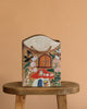 The Odin Parker x Gemma Koomen Gift Bag, featuring a whimsical house shape and hand-painted florals, birds, and doors, rests on a round wooden stool against a plain beige background.