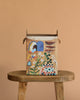 The Odin Parker x Gemma Koomen Gift Bag, adorned with vibrant artwork of a woman, birds, flowers, a blue purse, and a checkered blanket, sits on a wooden stool against a tan background—an eco-friendly choice for gifting.