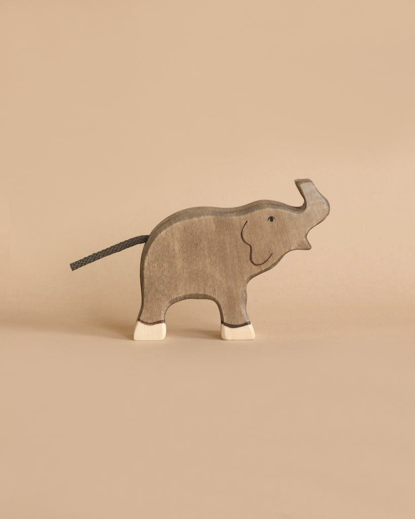 A Holztiger Handmade Wooden Safari Animals Set elephant with white feet and a black string tail stands on a beige surface, perfect for imaginative play or adding to your animal figures collection.