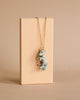 The Galaxy Unicorn Necklace, featuring a small, light blue unicorn pendant on a gold chain, is elegantly displayed against a light beige rectangular stand. This piece of whimsical wearables captures the imagination with its charming design and neutral background.