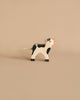 A small wooden toy cow with black and white patches stands on a plain beige background. Handcrafted in Europe, this Holztiger Black Calf figure features simple, minimalist details, including a short, rope-like tail and an orange snout.