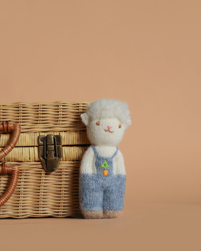 The Organic Alpaca Wool Doll - Sheep is a small, handmade plush with white fur, wearing blue overalls and holding a carrot, displayed next to a wicker basket on a plain beige background.