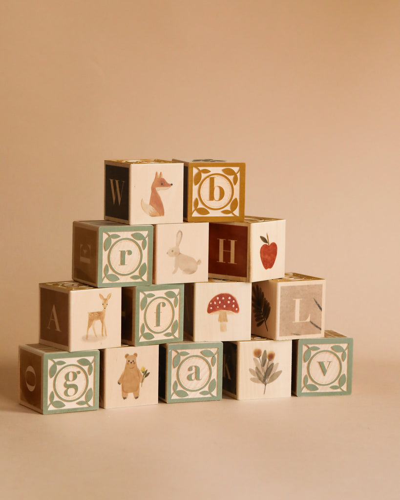 The Newborn Gift Set includes a pyramid of wooden alphabet blocks featuring letters and nature-themed illustrations—fox, rabbit, deer, bear, mushroom, flower, and strawberry—on a plain beige background.
