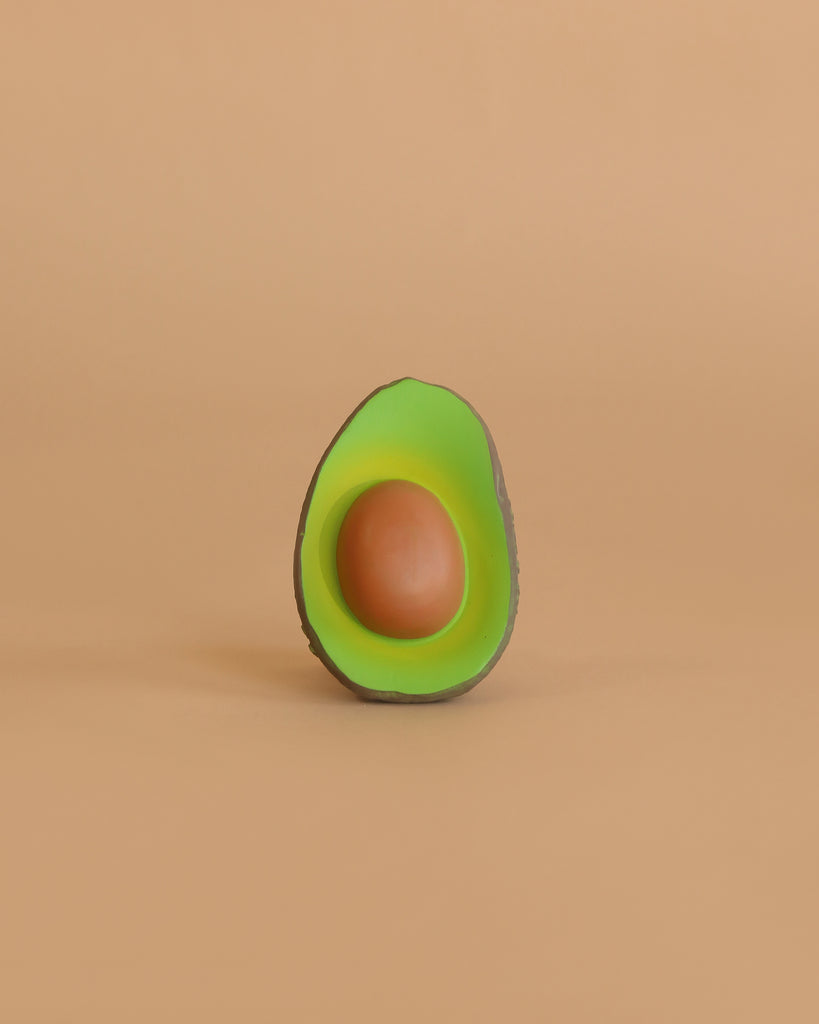 Arnold the Avocado Teething Toy, shaped like a halved avocado with green flesh and a brown seed, sits centered on a solid beige background—its natural rubber design makes it perfect for sensory play.