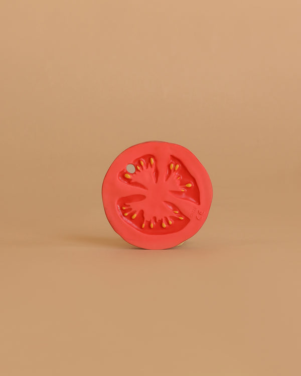 A vibrant red Renato the Tomato Teething Toy, crafted from natural rubber, stands upright against a plain beige background, with its detailed segments and seeds highlighted in the minimalist setting.