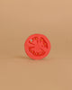 A vibrant red Renato the Tomato Teething Toy, crafted from natural rubber, stands upright against a plain beige background, with its detailed segments and seeds highlighted in the minimalist setting.