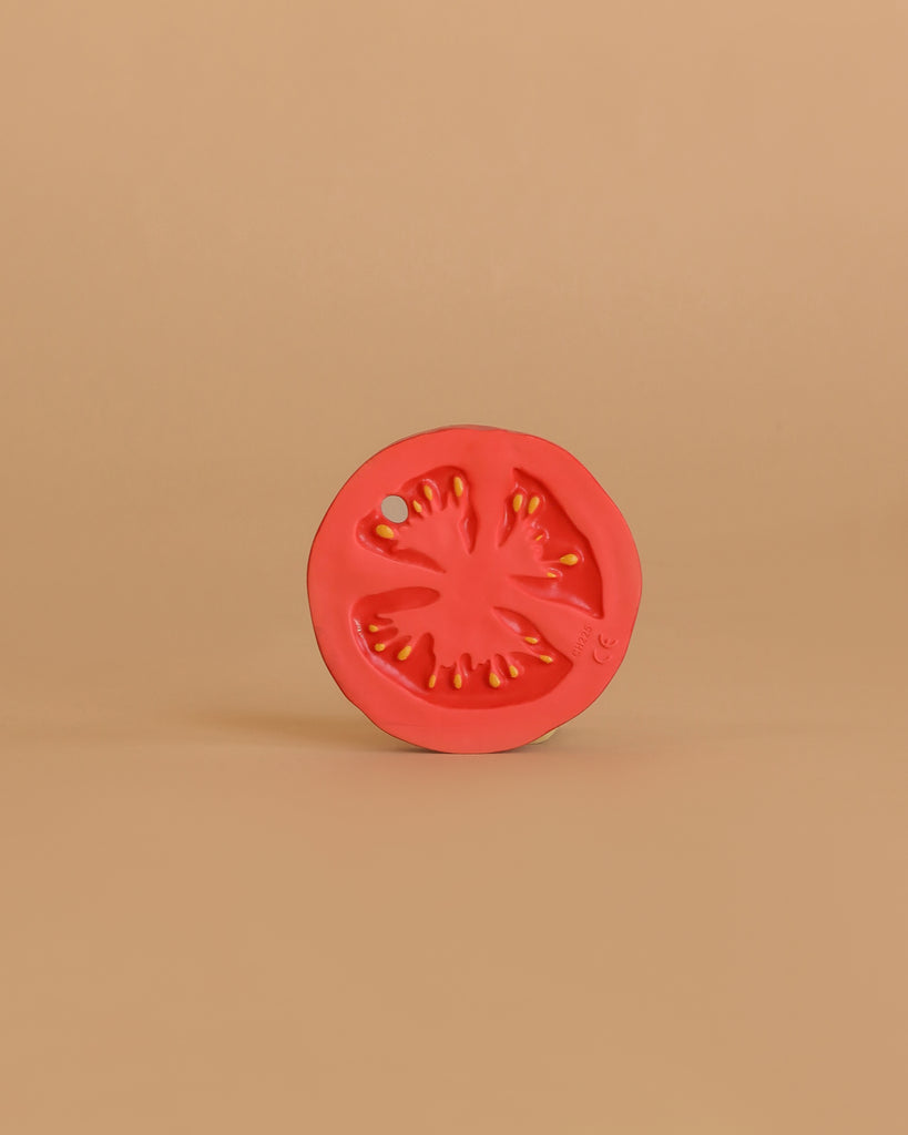 A vibrant red Renato the Tomato Teething Toy, crafted from natural rubber, stands upright against a plain beige background, with its detailed segments and seeds highlighted in the minimalist setting.