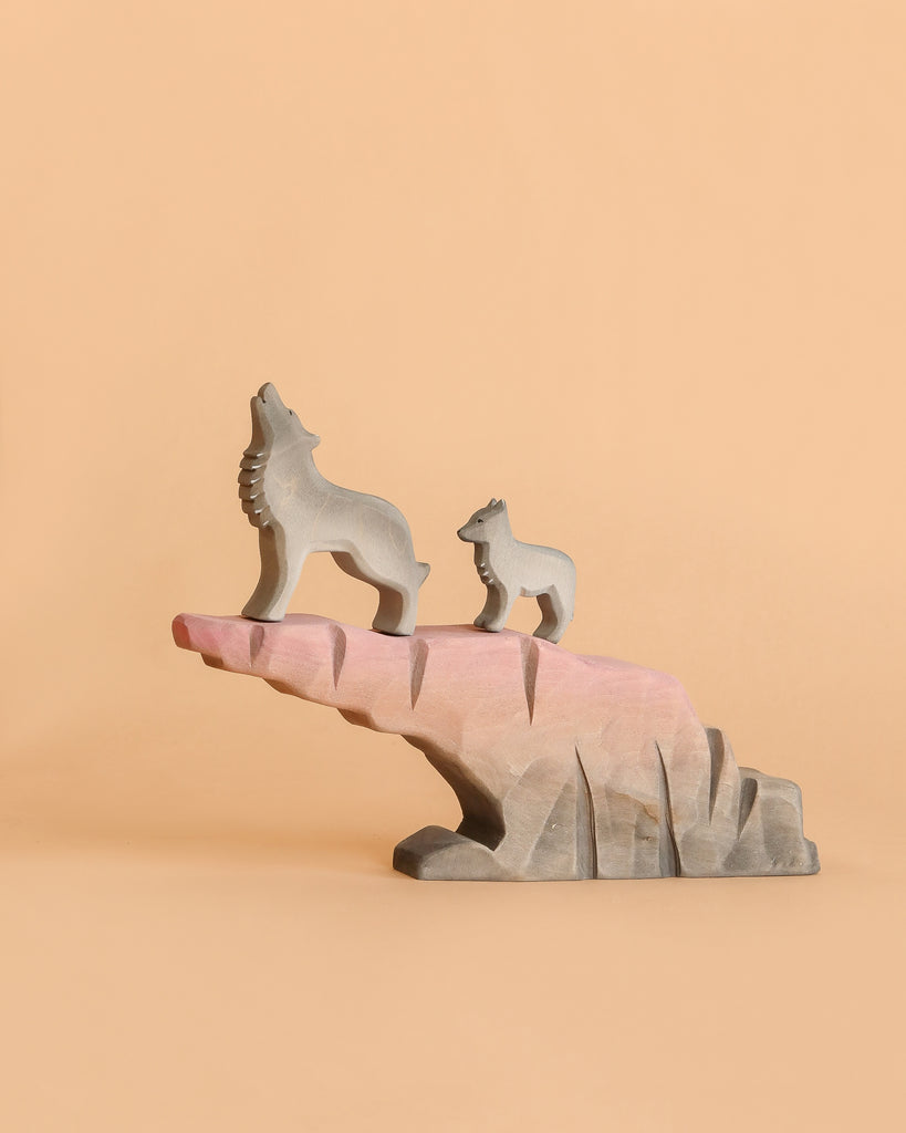 Two handcrafted wooden wolves from the Bumbu Howling Wolf Set stand on a pink and gray rock formation against a peach background. The larger wolf is howling while the smaller one stands beside it, both facing left.