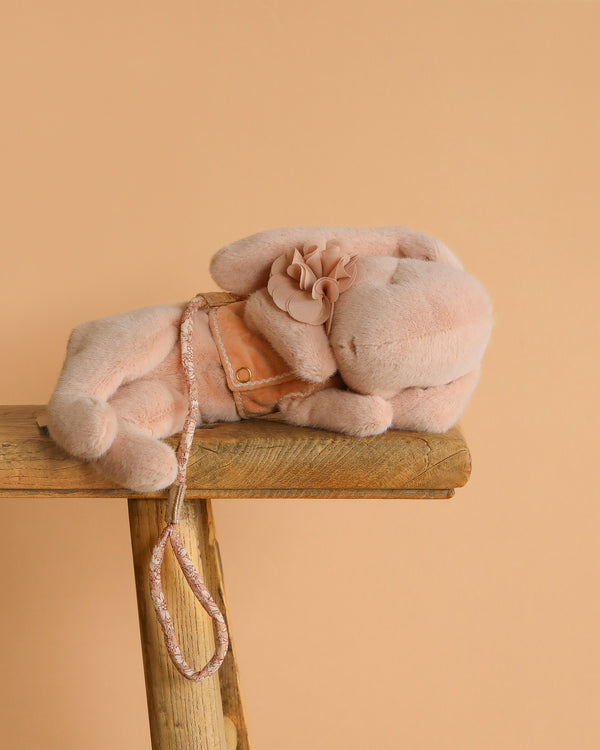 A Maileg Plush Sleeping Bunny With Harness in soft pink, featuring a flower on its ear and a matching harness, rests on a rustic wooden bench against a solid peach background.