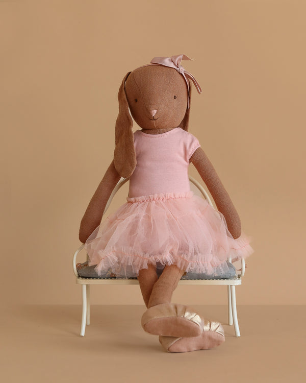 A Maileg Bunny, Size 3 in chocolate brown, dressed in a rose ballet suit and skirt, sits cross-legged on a small white bench against a beige background.