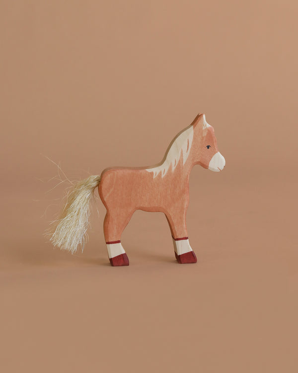 A handcrafted Holztiger Foal, Standing features a light brown body, white mane and tail, and red leg accents. Made in Europe with painted details, this wooden toy stands on a neutral beige background.