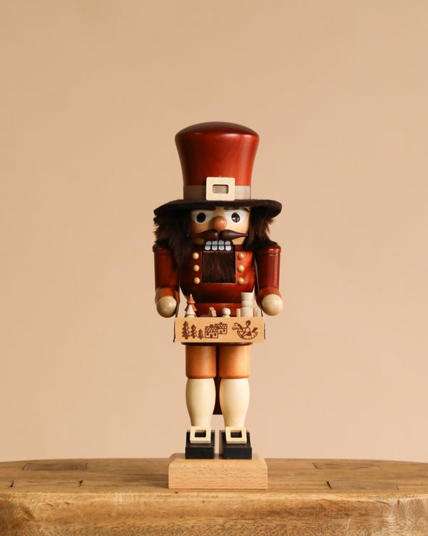 The Christian Ulbricht Collectible Wooden Nutcracker - Toy Trader features a tall brown hat, bushy beard, red coat, and beige shorts. It stands on a wooden surface against a beige background, holding a small decorated box.