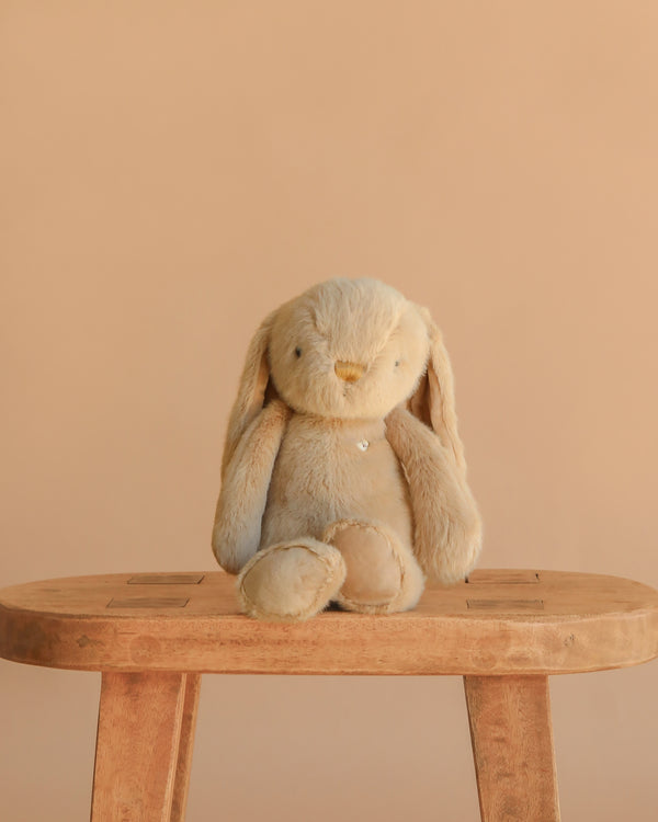 Snuggle Bunnies - Penelope - Caramel, a beige bunny with long ears, sits upright on a wooden stool against a plain light brown background.