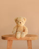 Snuggle Bunnies - Arthur the Bear, a light brown teddy with cream-colored paws and super soft plush fabric, sits on a wooden bench against a plain beige background.
