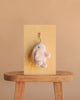 The Snuggle Bunnies - Penelope Keyring - Blossom, a soft light pink plush bunny, hangs on a hook attached to a wooden board set upright on a simple stool against a plain beige background.