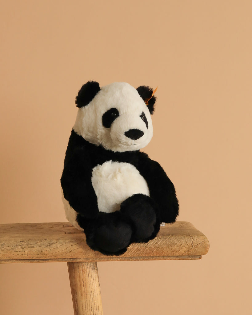 The Steiff Ming Panda Stuffed Animal - 11", featuring soft black and white fur, sits on a wooden stool against a plain beige background.