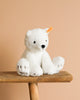 The Steiff Lasse Polar Bear Stuffed Animal - 11", crafted from ultra-soft white fabric and featuring a yellow ear tag, sits on a wooden bench against a plain beige background.