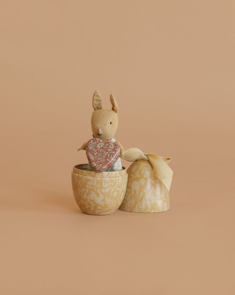 The Maileg Easter Egg with Micro Rabbit & Heart Set includes a micro rabbit holding a heart, nestled inside a decorative yellow egg with lid and ribbon, all pictured against a plain beige background.