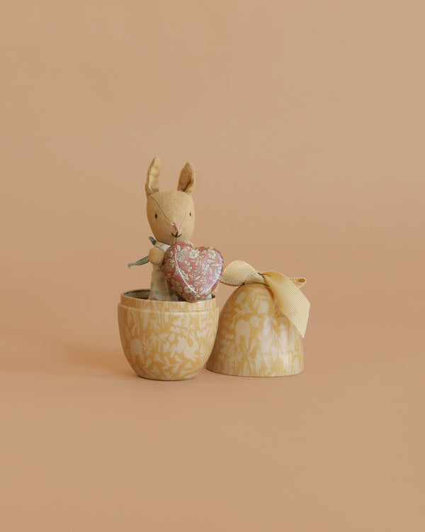 The Maileg Easter Egg with Micro Rabbit & Heart Set features a small stuffed bunny holding a pink heart inside an egg-shaped container with lid and yellow ribbon, displayed against a plain beige background.