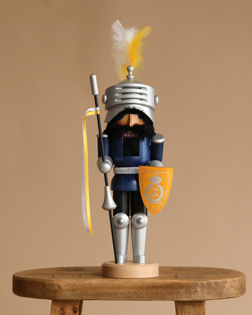 The Christian Ulbricht Collectible Wooden Nutcracker - Knight Lancelot Glazed stands on a stool in blue armor, helmet with yellow and white feather plume, and holds a yellow shield and lance—an eye-catching collectible.