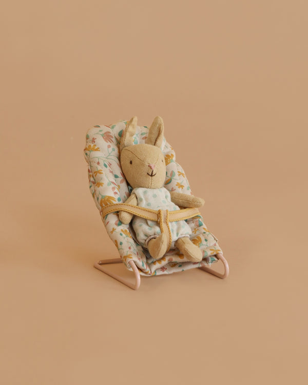 A Maileg Little Rabbit’s Nursery Set features a rabbit in a green dress strapped into a floral baby bouncer, mirroring the cozy comfort of the nursery set, all against a plain beige background.