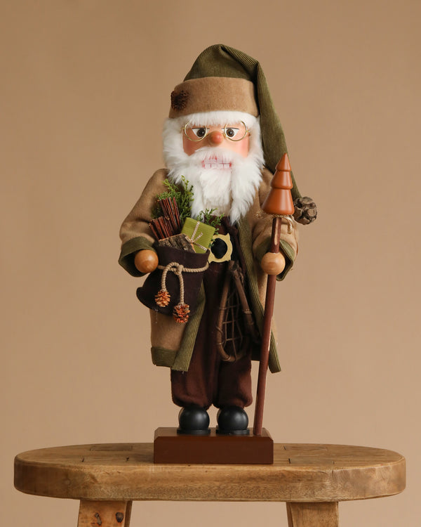 The Christian Ulbricht Collectible Wooden Nutcracker - Forest Santa Limited Edition 2500 holds a staff and a gift sack, standing on a wooden stool against a plain beige background.
