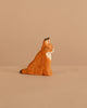 The Ostheimer Lynx Sitting, a small handcrafted wooden figurine painted orange with brown spots and a white chest, perfect for imaginative play, sits on a beige surface against a matching beige background.