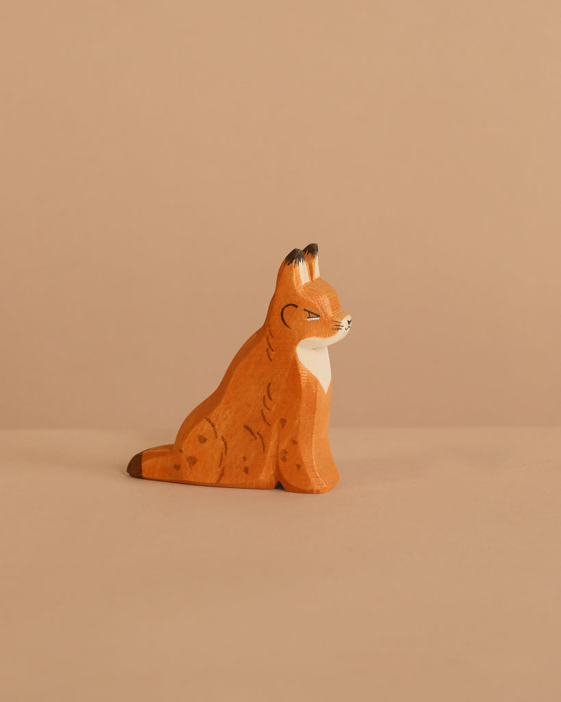 The Ostheimer Lynx Sitting, a small handcrafted wooden figurine painted orange with brown spots and a white chest, perfect for imaginative play, sits on a beige surface against a matching beige background.
