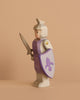 The Ostheimer Blue Knight - Standing is a handcrafted wooden toy with a light blue body, holding a sword and a shield adorned with a blue fleur-de-lis—ideal for imaginative play against any simple backdrop.