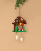 The Christian Ulbricht Cuckoo Clock Ornament, handcrafted in Germany, hangs by a red string. Shaped like a small house with brown roof, green trees, yellow bird, and red mushroom accents, it has cone-shaped weights and small parts that may pose a choking hazard.